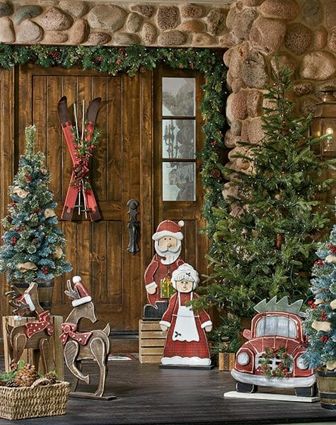 Stone house with a wood front door hung with skis, and wood Christmas cutouts by lit trees.