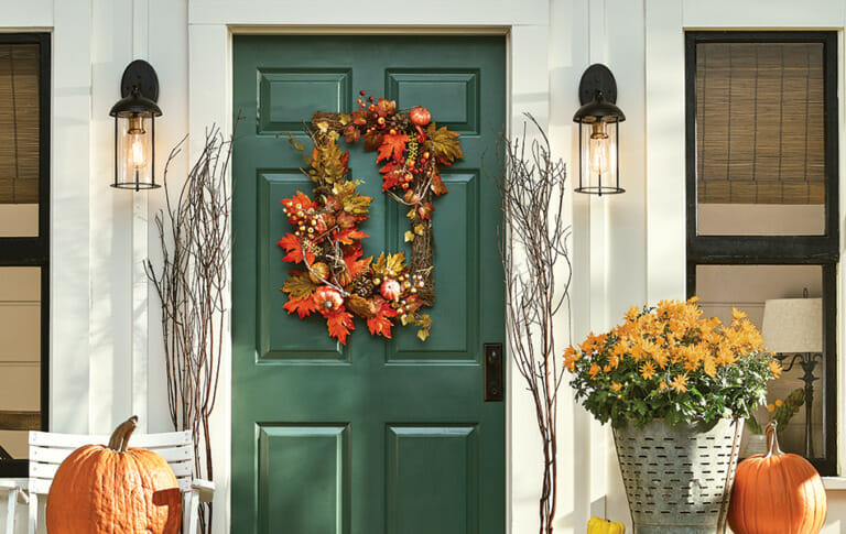 Green front door with rectangular Fall leaves twig wreath, lit lanterns, pumpkins, and pierced tin planter with gold mums.