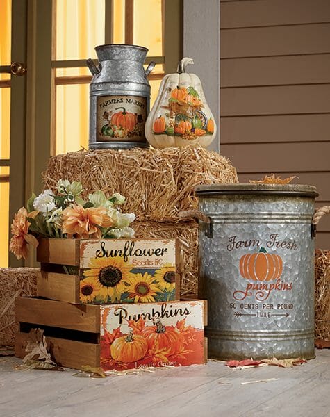Stacked straw bales on a porch with galvanized metal cans, painted white pumpkin, and two wood crates with flowers.