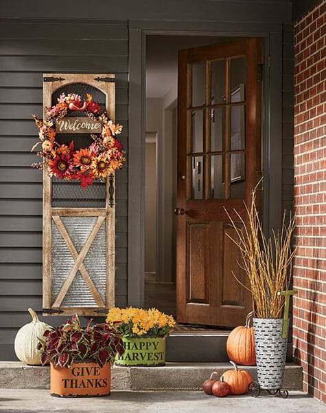 Harvest metal planters with coleus and mums, a shutter with a sunflower wreath, and a galvanized olive bucket with twigs.