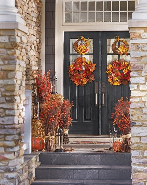 Double front doors with four Fall wreaths, four stick bundles with bittersweet, lit lanterns, and Welcome Friends pumpkins.