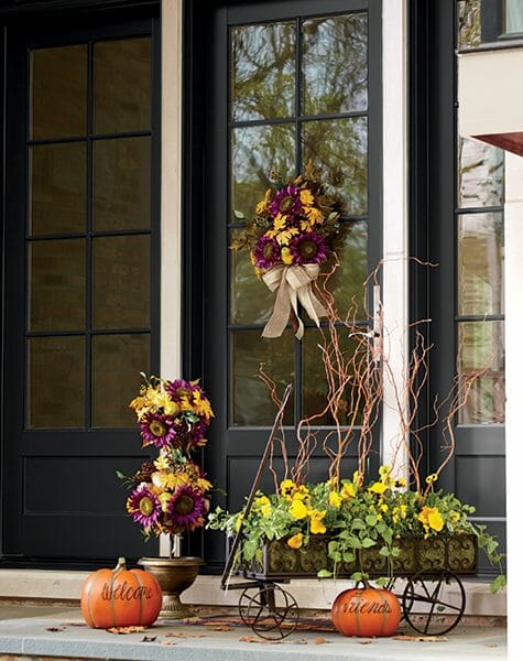 Fall purple sunflower topiary and a wreath on the front door, a metal cart with yellow pansies and twigs, and two pumpkins.