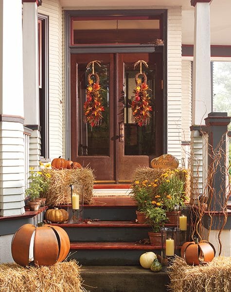 Two Fall swags on French doors, straw bales with metal pumpkins and gold mums, and lit candles in glass candleholders.