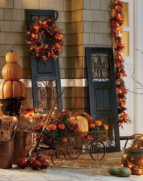 Lit Fall garlands and a wreath on black scroll shutters, and a metal cart filled with orange mums and pumpkins.
