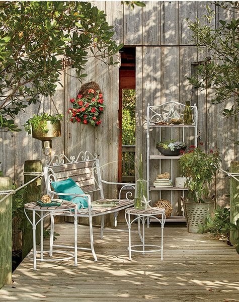A Farmhouse patio in front of a barn, with white scroll bench and shelving, hanging baskets and flowers.