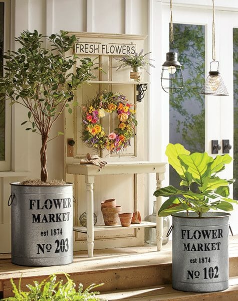 Farmhouse porch with galvanized buckets with plants, a potting bench with a wreath, and hanging wire cage lights.