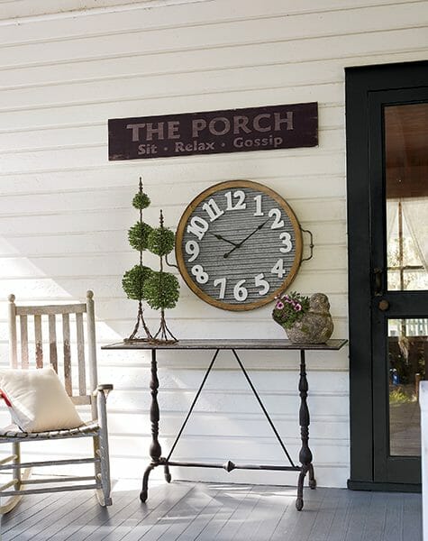 Farmhouse porch with whitewash rocker, turned leg table with topiaries, large round clock, black screen door and PORCH sign.