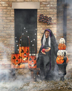 Halloween witch with an apple, three lit metal Jack-o-Lanterns and a BOO marquee, and a pumpkin topiary.