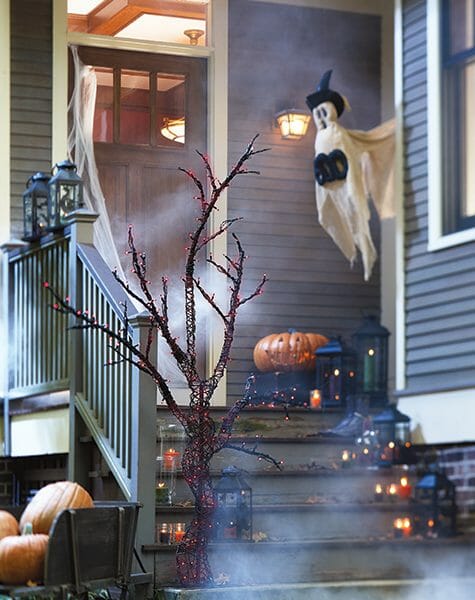 Halloween porch with lit black lanterns, lit Jack-o-Lantern, orange lit tree branch, and hanging white ghost that says BOO.