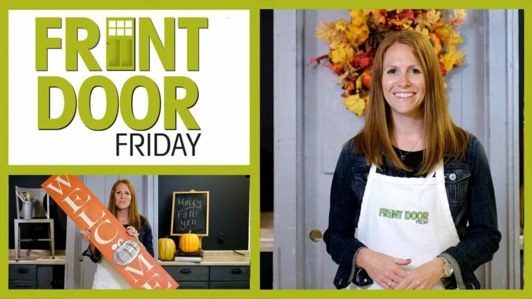 Front Door Friday – Smiling woman in an apron by a Fall wreath, and holding a tall orange Welcome sign by pumpkins.