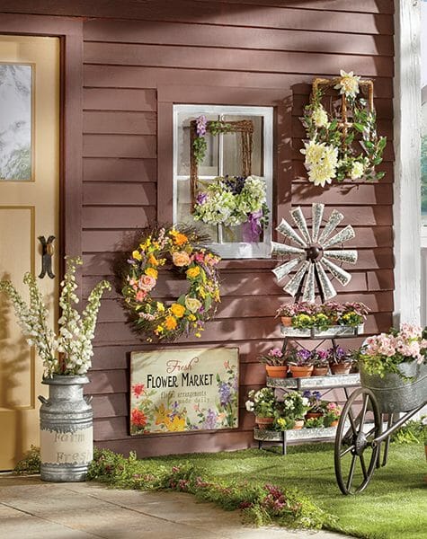 Outdoor gallery wall with displayed wreaths, a tiered windmill shelf unit with potted flowers, and a milk can of delphiniums.