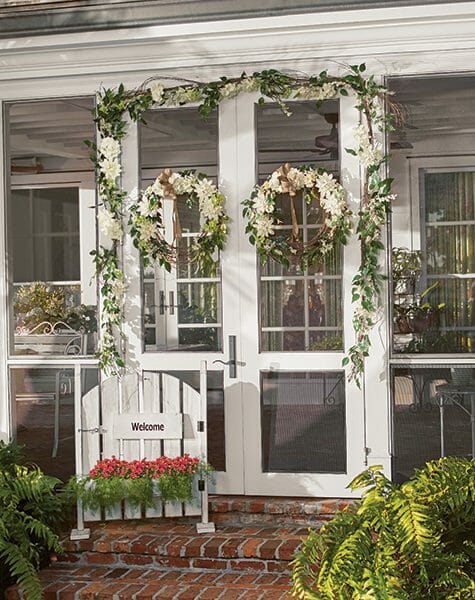 White screened porch double doors with two white floral wreaths and garlands, and a white gate planter with red flowers.