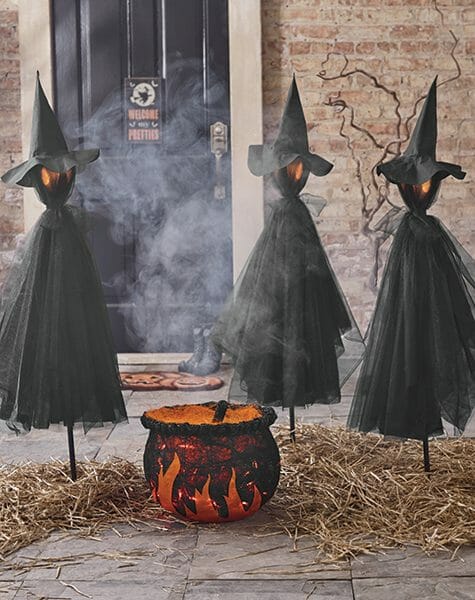 Three lit witch stakes with a lit cauldron, a Jack-o-Lantern doormat, and a Halloween sign on the black front door.