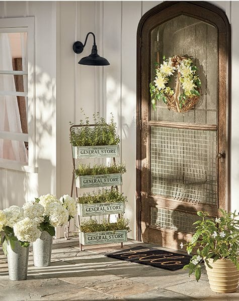 Farmhouse wood screen door with white floral wreath, tiered plant stand filled with herbs, and white hydrangeas in buckets.
