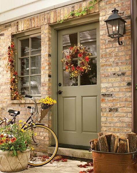 Fall wreath on a green front door, a bike basket holding yellow mums, and an antique copper boiler holding firewood.