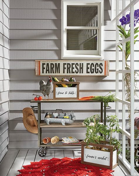 Farm Fresh Eggs sign on a porch with a cart holding squash, carrots, tomatoes, and a chicken figurine.