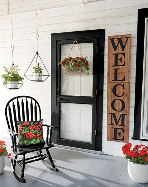 Farmhouse porch with a black rocker and red geranium pillow, red geraniums in planters and baskets, and a tall WELCOME sign.