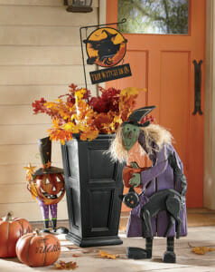 Orange front door with a tall black planter filled with Fall leaves, a wood witch cutout, and a metal Jack-o-Lantern.