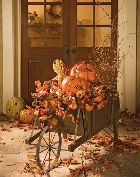 A wood wheelbarrow filled with Fall leaves and Welcome Friends pumpkins, in front of wood double doors.