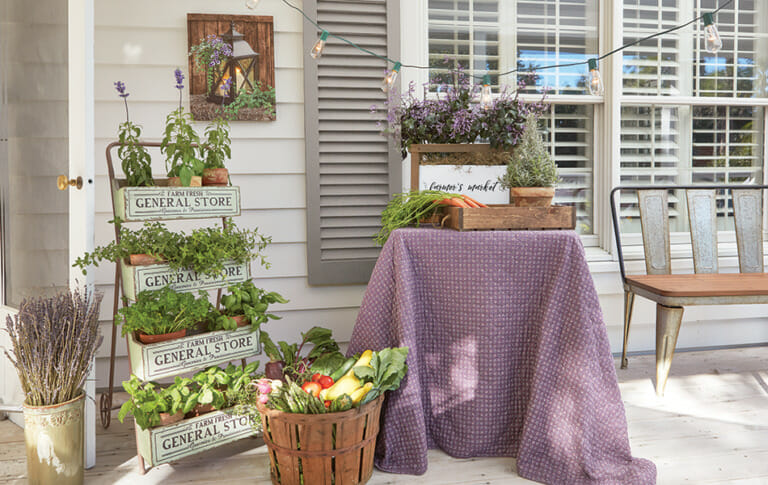 A Farmhouse porch with a tiered herb planter, a bushel basket of vegetables, a planter of purple flowers on a metal table.