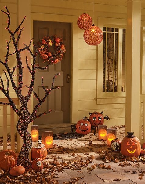 Lit Jack-o-Lanterns, luminaries, a tree, and hanging orange globes, and a Fall wreath on a brown front door.