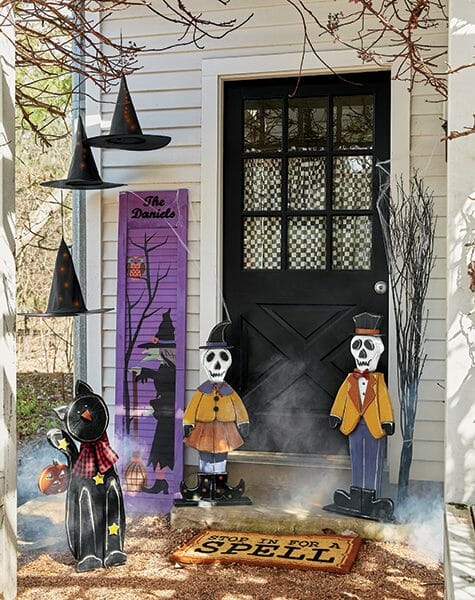Hanging witch hats, wood skeleton couple and black cat cutouts, and a purple witch shutter by a black front door. 