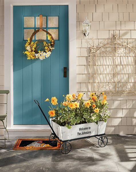 Teal door with forsythia wreath, a chicken with chicks doormat, and a personalized white wagon filled with yellow flowers.