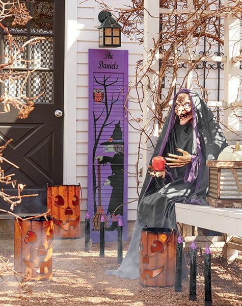 A witch holding an apple by a black front door, lit metal Jack-o-Lanterns, and a purple shutter with a witch painted on it.