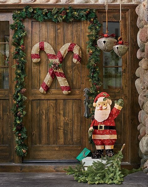 Candy cane hanger on a wood plank door with garland, two large hanging jingle bells, and a wood cutout Santa.