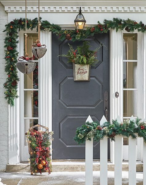 Gray front door with galvanized basket of greens, holiday garland,  large hanging jingle bells, and a decorated sleigh.