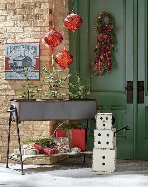 Green doors with a Winter swag, three large red ornaments, and a tin planter with pine trees and wrapped presents.
