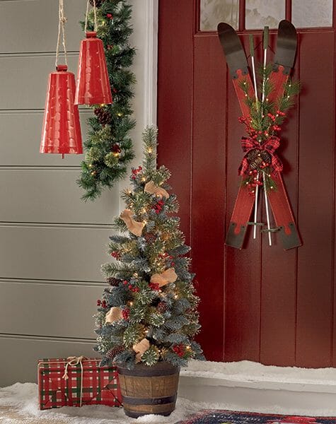 Front porch with two crossed skis with pine and bow hung on a red door, two red bells, and a small Christmas tree.